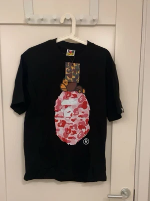 Svart Bape t-shirt med camomönster - Svart t-shirt från Bape med ikoniskt camomönstrat tryck i rosa och rött framtill samt en apa ovanpå. Klassisk passform och rund halsringning. Perfekt för dig som gillar streetwear och vill sticka ut med en unik design.