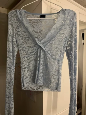 Ljusblå spetsblus med v-ringning - Säljer en ljusblå, transparent spetsblus med v-ringning och långa ärmar från Gina tricot. Blusen har ett omlottliknande fall framtill och är perfekt för dig som gillar romantiska detaljer och en feminin look.