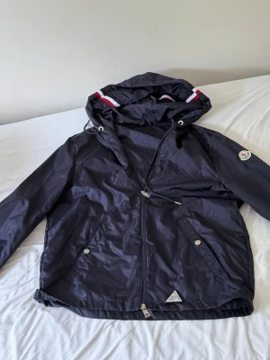 Mörkblå vindjacka från Moncler - Snygg mörkblå vindjacka från Moncler med huva och dragkedja. Jackan har vita och röda detaljer på huvan, två fickor med tryckknappar och Moncler-logga på ärmen. Tillverkad i lätt och glansigt material som står emot vind.