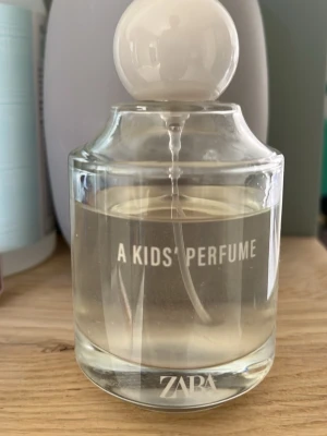 A Kids Perfume från ZARA - En stilren parfymflaska från ZARA med texten 'A KIDS PERFUME' på framsidan. Flaskan är genomskinlig med rund vit kork och innehåller en ljusgul vätska. Perfekt för dig som gillar minimalistisk design och fräscha dofter.