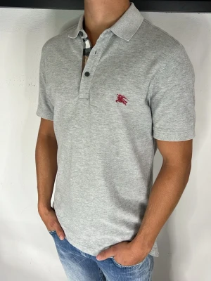 Burberry Piké  - Snygg grå pikétröja i bomull med klassisk krage och knappslå. Tröjan har korta ärmar och en liten röd broderad logga på bröstet. Perfekt till jeans för en clean och enkel stil.