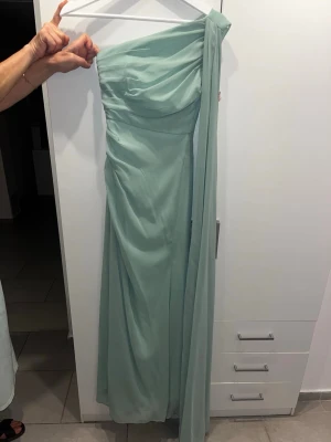 Ljusgrön långklänning oneshoulder - Elegant ljusgrön långklänning i flowigt material med oneshoulder-design och draperad överdel. Klänningen har en lång, svepande detalj från axeln och en figurnära passform upptill. Perfekt för speciella tillfällen. Använd en gång och köpt för 1500kr. för bal och bröllop etc. Midjan är professionellt omsydd tightare. så perfekt för dig som har smal midja. 