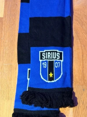 Sirius halsduk blå och svart - Säljer en snygg halsduk med Sirius klubbmärke och årtalet 1907. Halsduken har breda blå och svarta ränder samt fransar i ändarna. Perfekt för dig som vill visa ditt stöd för Sirius på matchen eller i vardagen.