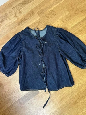 Mörkblå jeansblus med knytband - Trendig mörkblå jeansblus i denim med puffiga trekvartsärmar och knytband framtill. Blusen har rund halsringning och en avslappnad passform som ger en cool och avslappnad vibe. Perfekt att styla med både kjol och jeans. Inga lappar kvar men helt oanvänd. 