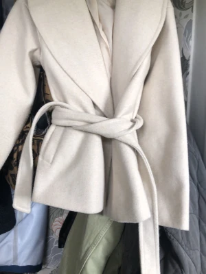 Beige kappa med knytskärp - Stilren beige kappa med bred krage och knytskärp i midjan. Kappan har raka ärmar och två sidofickor. Tillverkad i ett mjukt tyg som ger en lyxig känsla och passar perfekt till lager-på-lager-stil.