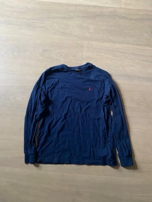 Mörkblå långärmad topp Polo Ralph Lauren - Mörkblå långärmad topp från Polo Ralph Lauren med rund halsringning och liten röd broderad logga på bröstet. Tillverkad i mjuk bomull som känns skön mot huden. Perfekt för en avslappnad och stilren look.