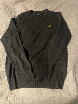 Mörkgrå tröja från Lyle & Scott - Mörkgrå stickad tröja från Lyle & Scott med rund halsringning och den klassiska gula logon broderad på bröstet. Tröjan har långa ärmar och ribbade muddar vid ärmslut och nederkant. Perfekt för lager på lager och stilren look.