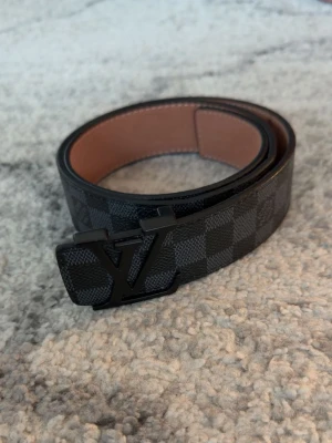 L V bälte - Snyggt svart bälte med rutigt mönster och ett stort spänne i metall. Bältet har en klassisk design och insidan är i beige. Perfekt accessoar för att lyfta din outfit.