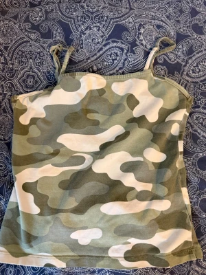 Camouflagefärgad linnetopp med knyt pratar pris - Säljer en ärmlös linnetopp med smala axelband och knyt på axlarna. Toppen har ett grönt, vitt och beige camouflage-mönster och är croppad i modellen. Perfekt för dig som gillar streetstyle och vill sticka ut.   PRATAR PRIS