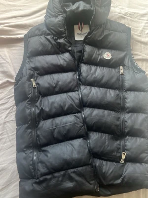 Svart dunväst från Moncler - Snygg svart dunväst från Moncler med två vertikala dragkedjefickor framtill och Moncler-logga på bröstet. Västen har hög krage och quiltad design som ger en sportig och trendig look. Perfekt för lager-på-lager och streetstyle.
