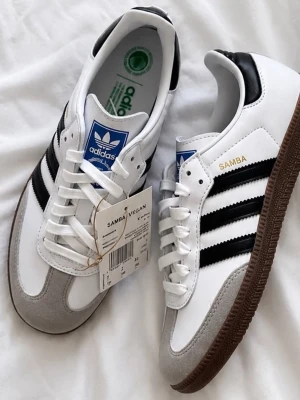 Adidas Samba Vegan vita sneakers - Klassiska Adidas Samba Vegan sneakers i vitt med svarta ränder och detaljer. Skorna har brun sula, blå logga på plösen och grå mockaliknande tåparti. Tillverkade i veganska material och har en retroinspirerad design med snörning.