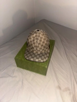 Beige keps från Gucci äkta!! - Säljer en beige keps från Gucci med det klassiska GG-monogrammönstret över hela kepsen. Kepsen har böjd skärm och detaljer i grönt och rött på sidan. Tillverkad i canvasmaterial och har en stilren och lyxig look. Box, kvitto med följer 