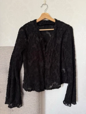 Svart transparent blus med knappar - Säljer en svart transparent blus med långa ärmar och knappar framtill. Materialet är lätt och har en rynkad struktur som ger en cool look. Perfekt att styla över ett linne eller topp för en trendig outfit.