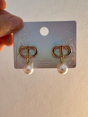 Guldfärgade örhängen med pärlor - Saljer ett par jättefina guldfärgade med hängande vit pärlor. Skick: Helt nya och oanvända (sitter kvar på displaykortet) Klassisk, feminin och lyxig design som passar till allt.