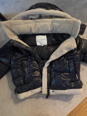 Svart pufferjacka med teddyfoder - Snygg svart pufferjacka med mysigt beige teddyfoder på insidan och i kragen. Jackan har dragkedja, knappar, två stora fickor framtill och justerbar midja. Luvan går att ta av genom dragkedjan. Perfekt för kalla vinterdagar och ger en cool look med sin glansiga yta och rejäla krage.