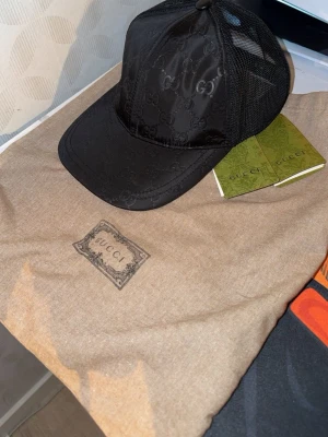 Svart keps från Gucci med GG-mönster - Snygg svart keps från Gucci med det klassiska GG-monogrammet över hela kepsen. Framsidan är i blankt tyg och baksidan har mesh för extra ventilation. Perfekt accessoar för en trendig och exklusiv look. !!Dust bag och tags ingår!! Helt ny!!