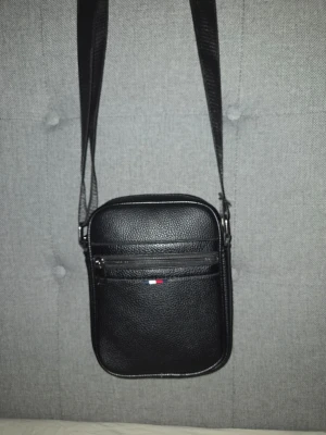 Svart tommy hilfiger axelväska - Snygg svart axelväska i skinnimitation med justerbar rem och flera fack med dragkedja. Liten logga framtill i rött, vitt och blått ger en stilren touch. Perfekt storlek för det viktigaste och enkel att matcha till olika outfits.