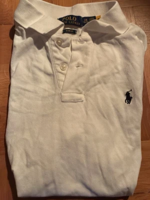 Vit pikétröja från Polo Ralph Lauren - Klassisk vit pikétröja från Polo Ralph Lauren med krage, knappar framtill och den ikoniska broderade loggan på bröstet. Perfekt till jeans eller shorts för en clean och stilren look.
