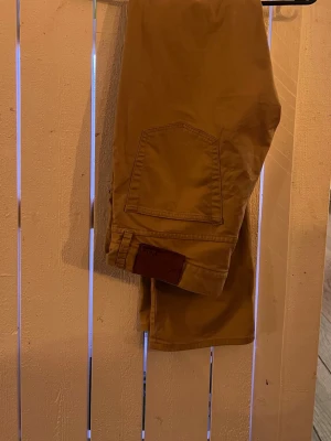 Beiga chinos från Polo Ralph Lauren - Snygga beiga chinos från Polo Ralph Lauren. Byxorna har framfickor, bakficka med knapp och detaljer i brunt läder vid linningen. Tillverkade i mjukt bomullstyg som känns bekvämt hela dagen
