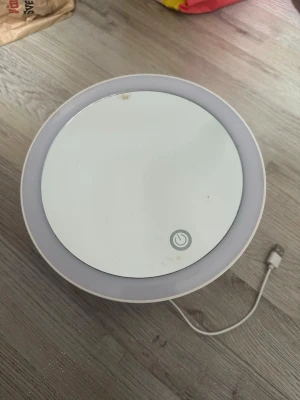 Rund sminkspegel med LED-belysning - Säljer en rund sminkspegel i vitt med inbyggd LED-belysning och touch-knapp på framsidan. Spegeln har USB-sladd för laddning och justerbart stativ på baksidan. Perfekt för sminkning och styling med jämnt ljus. Man kan sätta fast den med klistret i bak