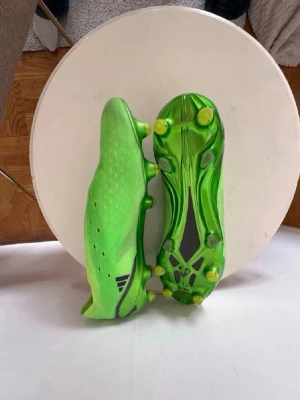 Gröna adidas fotbollsskor - Säljer ett par grymt snygga fotbollsskor i neon-grönt med svarta detaljer och X-mönster på hälen. Saknar snörning, då dem används bättre utan. Skorna har låg profil, slitstark ovandel i syntetmaterial och fasta dobbar för spel på gräs. Perfekta för dig som vill sticka ut på planen!