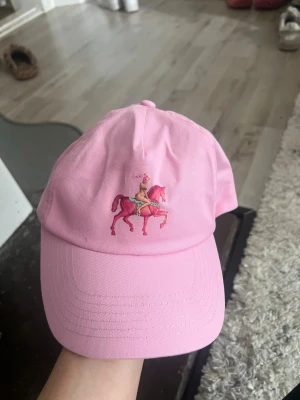 Rosa keps med broderad ryttare - Snygg rosa keps med böjd skärm och broderad detalj framtill som föreställer fröken snusk. Kepsen har klassisk passform och justerbar rem bak. Tillverkad i bomullstyg som känns lätt och bekvämt. Båda för 50kr