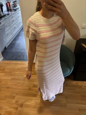 Randig stickad långklänning - Säljer en stickad långklänning med korta ärmar och rund hals. Klänningen har ett randigt mönster i vitt och ljusrosa och är rak i modellen med slits nertill. Öppen rygg med rosett. Aldrig använd, prislappen är kvar. 