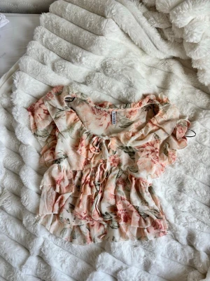 Blommig volangblus - Söt blommig blus med volanger och snörning framtill. Toppen har korta puffärmar och ett romantiskt mönster i rosa, grönt och beige. Tillverkad i ett lätt, luftigt material som passar perfekt till varma dagar.