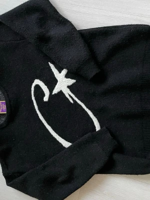 Svart stickad tröja med stjärnmotiv - Säljer en svart stickad tröja med ett vitt stjärnmotiv och böjd linje på framsidan. Tröjan har rund halsringning och långa ärmar. Materialet ser ut att vara mjukt och mysigt, perfekt för kyligare dagar.