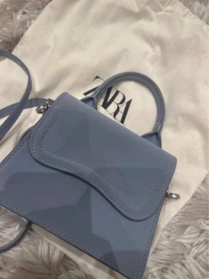Ljusblå minihandväska från Zara - Trendig ljusblå minihandväska från Zara med avtagbar axelrem och topphandtag. Väskan har en stilren, fyrkantig form och slät yta i konstläder. Perfekt accessoar för att lyfta din outfit.
