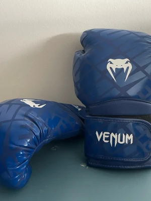 Blå boxningshandskar från Venum - Säljer ett par blå boxningshandskar från Venum med vitt logotryck och coolt mönster. Handskarna har kardborreknäppning runt handleden för extra stöd och är tillverkade i slitstarkt syntetmaterial. Perfekta för träning eller sparring.