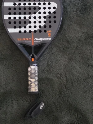Bullpadel padelracket svart/grå - Säljer ett Bullpadel padelracket i svart och grått med stora vita detaljer och orange inslag. Racketen har perforerad träffyta och grepp med mönstrad linda samt handledsrem. Perfekt för dig som vill ta ditt padelspel till nästa nivå.