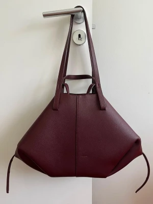 Vinröd shoppingväska i läder - Stilren shoppingväska i vinrött läder med unik geometrisk form och långa axelband. Väskan har minimalistisk design och små diskreta detaljer, perfekt för dig som gillar trendiga accessoarer med mycket plats.