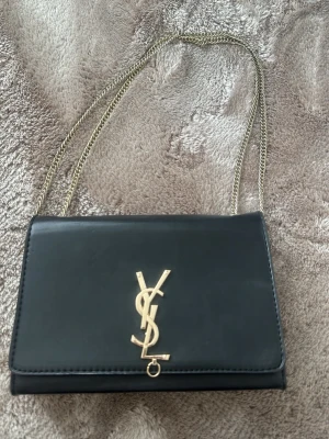 Svart axelremsväska med guldkedja - Snygg svart handväska från Yves Saint Laurent med guldfärgad kedjerem och ikonisk YSL-logga framtill. Väskan har en stilren, rektangulär form och är tillverkad i slätt läder. Perfekt för dig som vill ha en klassisk och trendig accessoar.