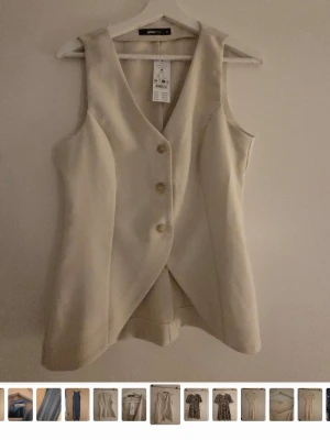 Beige lång väst med knappar - Stilren beige väst med tre knappar framtill och längre passform. Västen har en omlottliknande front och är ärmlös med djup v-ringning. Perfekt att styla över en skjorta eller t-shirt för en trendig look.