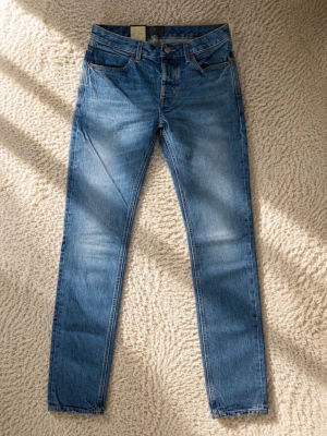 Neuw Stockholm jeans  - Säljer ett par blå jeans med smal passform och ett unikt svartvitt tryck på ena bakfickan. Jeansen har klassisk femficksdesign, knappgylf och är tillverkade i denim. Perfekta för dig som vill ha något som sticker ut lite extra.