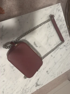 Vinröd axelväska med kedjerem - Snygg vinröd axelväska från Guess i fyrkantig modell. Väskan har en silverfärgad kedjerem med matchande vinrött axelparti och dragkedja upptill. Tillverkad i ett strukturerat konstläder som ger en lyxig känsla. Perfekt för att lyfta din outfit.