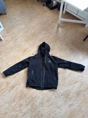 Svart vindjacka från Arc'teryx - Snygg svart vindjacka från Arc'teryx med huva och dragkedja framtill. Jackan har två snedställda fickor med dragkedja och logga på bröstet. Materialet är tunt och vindtätt, perfekt för blåsiga dagar.