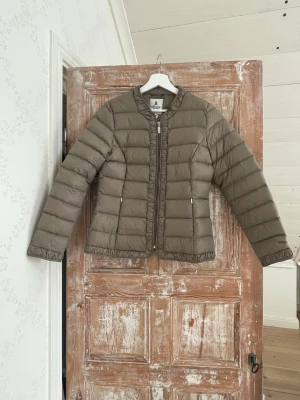 Beige dunjacka med dragkedja - Snygg beige dunjacka med quiltad design och rund halsringning. Jackan har dragkedja framtill, två fickor med dragkedja och diskreta detaljer längs kanterna. Perfekt för  hösten och enkel att matcha med olika outfits.