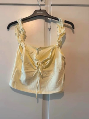 Gul croppad linnetopp med spets - Söt ljusgul croppad linnetopp med spetsdetaljer på axelbanden och knyt framtill. Toppen har en feminin v-ringning och ärmlös design, perfekt för varma dagar. Materialet känns mjukt och lätt mot huden. Skick: har en miten märke under armen, ej testat få bort själv endast tvättat 