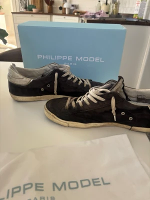 Philippe Model svarta låga sneakers - Snygga svarta sneakers från Philippe Model med vita detaljer och vita skosnören. Skorna har en låg siluett och är tillverkade i mocka och läder. Yttersulan är vit med en något sliten look och hälen har en silverfärgad detalj.