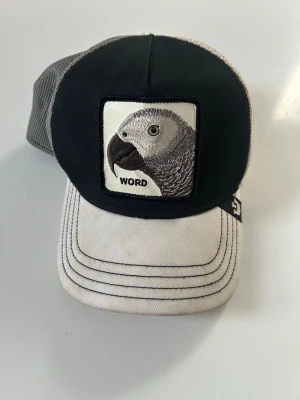 Svart och vit keps med papegojmärke - Snygg keps i svart och vitt med mesh baktill och broderad papegoja framtill där det står 'WORD'. Justerbar passform och böjd skärm. Perfekt för dig som vill sticka ut med en unik accessoar.