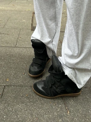 Svarta höga sneakers med kardborre - Säljer ett par svarta sneakers med högt skaft och breda kardborreband. Skorna har en chunky sula i brunt gummi och detaljer i både mocka och läder. Perfekta för dig som gillar streetwear och vill sticka ut med en unik look.               Kopior!!