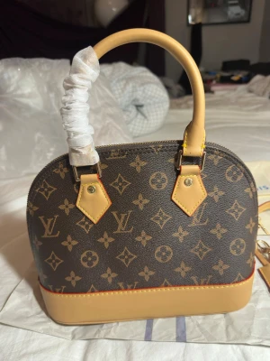 Louis Vuitton Monogram handväska - Snygg handväska från Louis Vuitton i klassiskt monogrammönster. Väskan har rundad form, bruna och beige färger, guldfärgade metalldetaljer och handtag i ljust läder. Invändigt finns en ficka och beige textilfoder. Perfekt för dig som gillar lyxiga accessoarer.