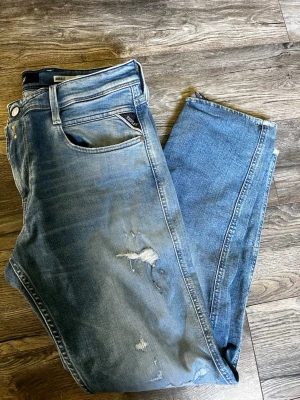 Blå slitna jeans  - Säljer ett par blå jeans  slitna detaljer på både lår och knän. Jeansen har klassisk femficksmodell och ljus tvätt med tydliga slitningar för en trendig look. de är väldigt bra i skicket aldrig använda . Det är en klassisk modell som heter anbass .Perfekta för dig som gillar en avslappnad och cool stil. 