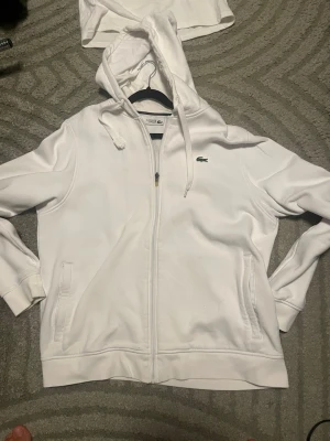 Vit hoodie med dragkedja från Lacoste - Säljer en clean vit hoodie från Lacoste med dragkedja framtill och klassisk krokodil-logga på bröstet. Hoodien har huva med dragsko, två fickor och ribbade muddar. Perfekt för en chill och sportig look.