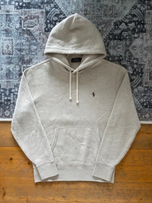 Beige hoodie från Polo Ralph Lauren - Snygg beige hoodie från Polo Ralph Lauren med klassisk huva och dragsnören. Har en broderad logga på bröstet och en stor magficka framtill. Tillverkad i mjukt bomullsmaterial och har ribbade muddar vid ärmar och nederkant.