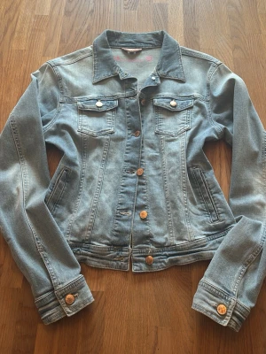Ljusblå klassisk jeansjacka - Säljer en ljusblå jeansjacka med snygga bronsfärgade knappar och två bröstfickor med lock. Jackan har en klassisk krage och raka ärmar med knapp vid ärmslut. Perfekt för att ge din outfit en cool touch.