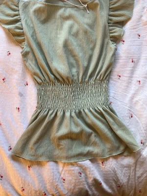 Ljusgrön ribbad blus med volang - Supersöt ljusgrön blus med ribbad struktur och volangärmar. Smockad midja ger en snygg passform och volang nertill gör toppen extra feminin. Perfekt att styla med jeans eller kjol för en trendig look.