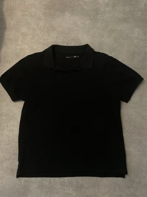 Svart pikétröja med krage - Snygg svart pikétröja med klassisk krage och knappar framtill. Tröjan är i enkel och stilren design, perfekt till jeans eller shorts. Materialet känns mjukt och bekvämt mot huden.